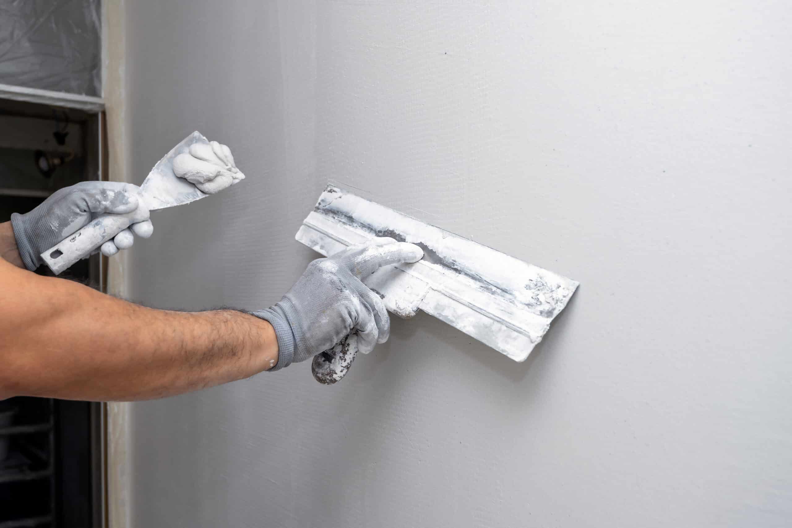 Behandschuhte Hände glätten eine Wand mit einem breiten Flächenspachtel.