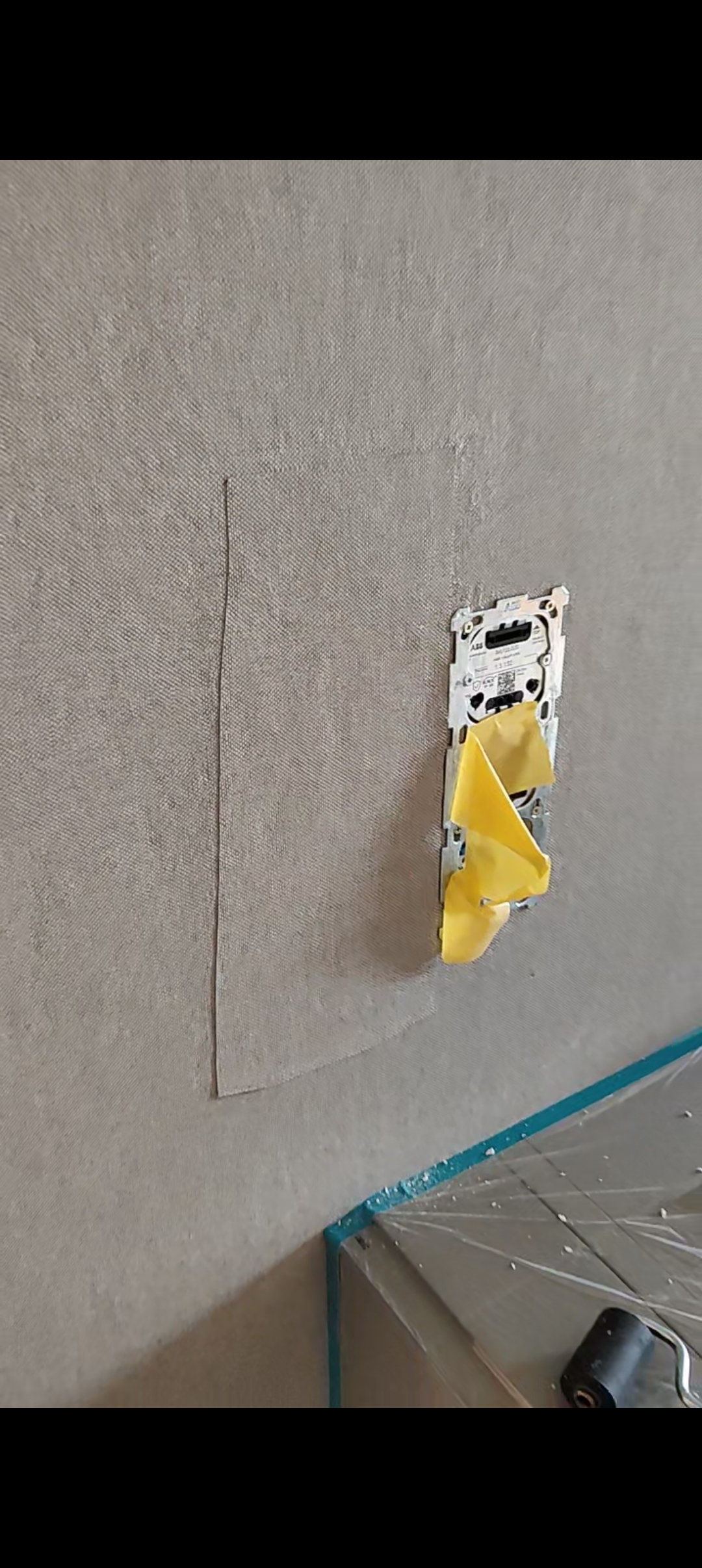Rechteckiges Tapetenstück auf einer grauen strukturierten Wand rund um eine abgedeckte Elektroinstallation.